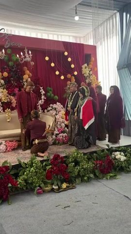 A wedding ceremony is taking place indoors. A man in a red outfit with traditional Indonesian headwear is standing, while another man in a similar outfit is kneeling before him, appearing to place something on his leg. Two other couples and a woman are standing to the right, observing the ceremony. The background features a red curtain, floral decorations in shades of red, pink, and yellow, and a chandelier. The floor is covered with a grey carpet and decorated with red and green floral arrangements.