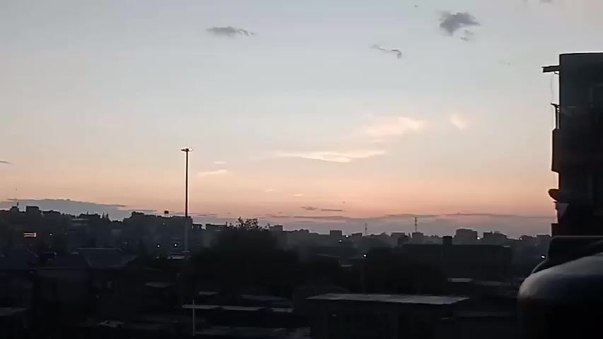 The video presents a skyline view of a city at dusk or dawn, with a light pink and grey sky. The cityscape is composed of buildings of varying heights, some appearing older with visible rooftops, and others looking more modern in the distance. A tall street lamp stands prominently in the midground, adding to the urban landscape. In the lower right corner, part of a building with a water tank on its roof is visible, framing the scene.  The overall atmosphere is calm and tranquil, captured during a time of transition in natural lighting.
