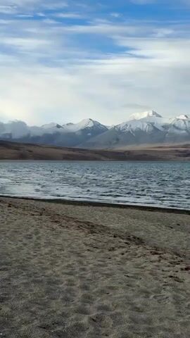 The video depicts a scenic view of a serene lake with snow-capped mountains in the background. The foreground shows a sandy beach leading to the water's edge, where small waves ripple across the lake's surface. The sky is partly cloudy, with patches of blue visible. The mountains are partially shrouded in clouds, and their peaks are covered in snow.