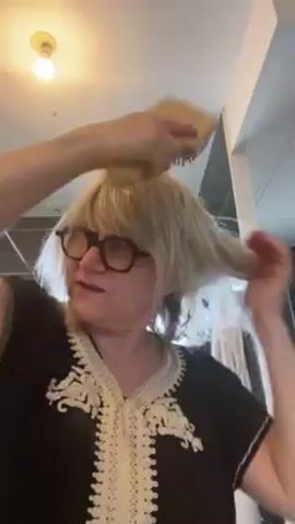 A woman with shoulder-length blonde hair and black-rimmed glasses is brushing her hair. She is wearing a black top with white embroidery. She looks directly at the camera with a slightly pouty expression. The background is a room with a light fixture and a white pillar.  The camera angle is low, looking up at the woman.