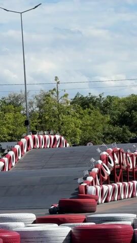 Two go-karts are racing up a ramp on a track. The go-karts have drivers wearing helmets. The track is bordered by tires painted red and white. Trees and a cloudy sky are visible in the background. A street light is also present.