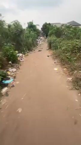 The video shows a first-person perspective of someone walking down a dirt path. The path is littered with trash, including plastic bags, wrappers, and other debris. The path is flanked by overgrown vegetation on both sides. In the distance, a building can be seen through the trees. The person is wearing sandals. The sky is overcast.