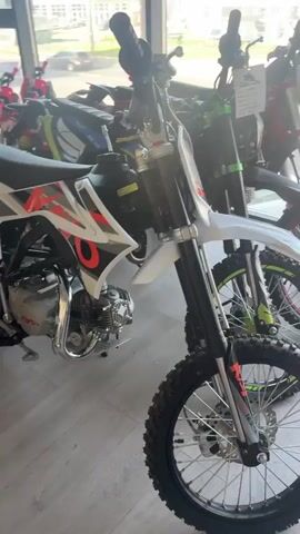 The video displays a row of dirt bikes parked inside a showroom. The bikes are lined up against a large window, through which a street scene is visible. The street appears to be in a town or city, with buildings, trees, and cars. The weather seems to be sunny, as indicated by the bright lighting. The time of day is likely daytime, possibly late morning or afternoon.

The dirt bikes are varied in color and design, with some being white, red, green, and black with yellow accents. They are all facing the same direction, with their front wheels pointed slightly to the left. The bikes have knobby tires, characteristic of off-road vehicles. Some of the bikes have price tags or other labels attached to them.

The foreground of the video focuses on the front wheel and suspension of a white dirt bike. The background shows the rest of the bikes in the row, as well as the street scene outside. The overall impression is one of a dealership showcasing its inventory of dirt bikes.

Visible text on the bikes includes brand names and model numbers. For example, one bike has "KAYO" printed on its side, and another has "SPARK" visible. Other text on the labels is too small to be legible. The general atmosphere is clean and well-lit, suggesting a professional retail environment. The scene conveys a sense of excitement and adventure associated with dirt biking.