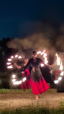Three individuals are performing a fire dancing routine at dusk. The dancers are wearing dark tops and flowing red and black skirts. They are holding flaming props, creating trails of fire as they move. The background shows a dark sky, trees, and what appears to be a body of water. The ground is paved with stones, and there is tall grass at the edges of the frame. Smoke from the fire is visible in the air.