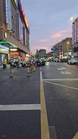A street scene at dusk with a pink and purple sky. On the left is a multi-story building with shops on the ground floor and illuminated signs. To the right of the shops, motorcycles are parked along the sidewalk. The street is paved with asphalt and marked with yellow and white lines. A crosswalk is visible in the foreground. Two women are crossing the street from right to left. The woman on the left is wearing a white layered dress and holding a phone. The woman on the right is wearing a striped long-sleeved shirt, jeans, and glasses, and is also holding a phone. Cars are visible on the street, and traffic lights are illuminated in the distance. The buildings on the right side of the street are modern in design.