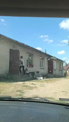 The video captures a rural scene in Zaręby Kościelne, Poland, on a clear, sunny day. The primary subject is a modest, single-story house with a gray facade and a sloping roof. In front of the house, a person, likely an elderly woman, sits on the steps leading to the entrance. She appears to be relaxed, perhaps enjoying the pleasant weather. Nearby, a bicycle and some other objects are visible, suggesting a lived-in environment. The yard is unpaved and appears somewhat dry, typical of a rural setting. The sky is bright blue with scattered white clouds, indicating fair weather. The overall atmosphere is peaceful and serene, reflecting a quiet moment in a rural Polish village. No text is visible in the video.