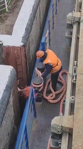 Here's a description of the video:

A person wearing an orange safety vest and hard hat is working with a thick, reddish-pink rope on a metal walkway. The person is standing between two dark-colored bollards and wrapping the rope around them. The walkway has a blue railing on one side. On the other side is a concrete and rusted metal wall. The background is slightly out of focus and shows vegetation and possibly another structure. The person is facing away from the camera. The video appears to have been taken from an elevated position.
