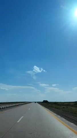 A first-person perspective video shows a vehicle traveling on a paved road under a clear blue sky. The sun is visible in the upper right corner, casting a bright glare. The road has a white dashed line in the left lane and a solid yellow line on the right shoulder. Vegetation and a fence line are visible on the right side of the road. A single vehicle is in the distance, ahead of the camera.