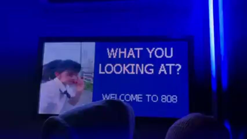 The video shows a rectangular screen that displays "WHAT YOU LOOKING AT?" and "WELCOME TO 808" in large white letters against a blue background. To the left of the text, there is a live video feed of a person in a white shirt who appears to be waving at the camera. The room is bathed in blue light, with neon light strips visible to the right of the screen. Two people are seen in the foreground, silhouetted against the blue light, wearing hats or hoods.