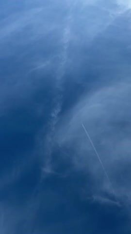 The video displays a clear blue sky with scattered wispy clouds. A jet plane is visible in the distance, leaving a prominent contrail that stretches across the frame diagonally from the lower right to the upper left. The contrail appears to be a straight line, indicating the plane's steady trajectory. Some fainter, feathery clouds are also present in the sky, adding depth and texture to the scene. The camera appears to be looking upwards from ground level.