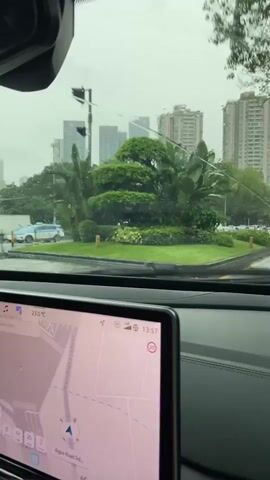 The video is shot from inside a vehicle, looking out through the windshield. A windshield wiper is visible on the left side of the frame, in motion. The weather appears to be rainy, with water streaking down the windshield. In the foreground, a large digital display is mounted on the dashboard, showing a map interface with the current time as 13:57. Outside the vehicle, there is a roundabout with well-maintained green landscaping, including trimmed bushes and plants. Beyond the roundabout, a street is visible with several lanes of traffic. Cars and at least one bus are driving on the wet road. In the background, several tall buildings rise into the overcast sky. The overall scene depicts a view from a vehicle driving in urban, rainy conditions.