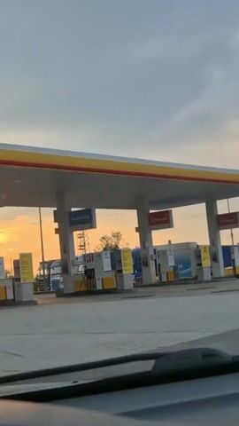 The video captures a view from inside a vehicle, looking out at a Shell gas station during what appears to be sunrise or sunset. The sky is filled with warm, golden light and some clouds. The gas station has a prominent yellow and red awning, with several pumps visible underneath. Signs on the pumps indicate "AutoGas" and "V-Power," with some pricing information visible on a yellow sign, including "Shell V-Power Racing 100," "V-Power Diesel," and "Shell FuelSave Diesel." The ground is paved asphalt. In the background, trees and power lines are visible, along with a truck. The overall mood is tranquil and serene, emphasized by the soft lighting of the sun.