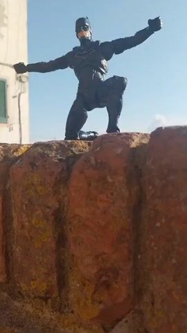A Batman action figure is posed on a stone wall, in front of a scenic landscape view of Peccioli, Italy. The action figure is dark gray and black, with a yellow bat symbol on its chest. It is in a kneeling position with its arms outstretched. The wall is made of rough, reddish-brown bricks and stones, with some patches of moss or lichen. In the background, there is a building with a white facade and a green shutter, a tall brick tower, and rolling hills dotted with houses and fields. The sky is clear and blue with a few scattered white clouds. The sun is shining brightly, suggesting it is daytime. The overall mood is dramatic and adventurous, fitting for the character of Batman.