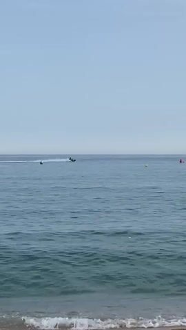 A static, eye-level shot from a beach shows a wide expanse of calm, blue ocean under a clear, pale blue sky. The shoreline, composed of sand and the edge of small waves, occupies the bottom of the frame. In the distance, two jet skis are visible on the water. One jet ski, positioned to the left, is a small dark silhouette. The other jet ski, to the right, is in motion, leaving a white wake behind it. A few small, indistinct buoys are scattered on the water's surface. The horizon line is clear, dividing the water from the sky.