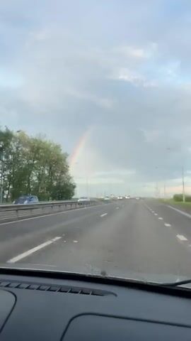 The video shows a view from the dashboard of a car driving on a multi-lane road. The road is wet and there is light traffic. A rainbow is visible in the sky ahead. The sky is mostly overcast. There are trees and light posts along the side of the road. A white bus is on the shoulder of the road.