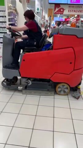 A person wearing overalls and a dark top is operating a large, red floor cleaning machine with a steering wheel and controls. The machine has gray and black components and yellow wheels. The person is seated and appears to be driving the machine down a tiled aisle in what looks like a large store or supermarket. Shelves stocked with products are visible on either side of the aisle, some labeled with "Perfumeria". Other shoppers and employees can be seen in the background, including a man in a light-colored shirt and dark pants walking away from the camera and a person on the far right, also walking. The store has a brightly lit ceiling with fluorescent lights and flags or decorations hanging from the ceiling. The floor is clean and tiled. The video appears to be shot from a slightly low angle and is in motion, following the cleaning machine.