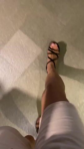 The video shows a person's legs and feet walking on a paved surface. The person is wearing white shorts and black sandals. The camera is positioned low and is moving, creating a blurry effect. The lighting suggests it is daytime or an area with artificial lighting. The paved surface appears to be made of large, rectangular stones. There are shadows visible on the ground, indicating a light source.
