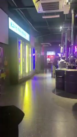The video shows the interior of a Korean BBQ restaurant. The floor is grey speckled concrete. Along the left wall is a long row of refrigerated cases, lit with yellow and blue vertical lights. A sign above them is in Korean. Tables are arranged in a row on the right, each with a metal grill and a metal ventilation pipe hanging overhead. The tables are made of dark gray metal barrels. There are people seated at the tables, eating. The back of one person’s head is visible in the lower left corner of the frame. The restaurant has a modern, industrial aesthetic with neon lighting.