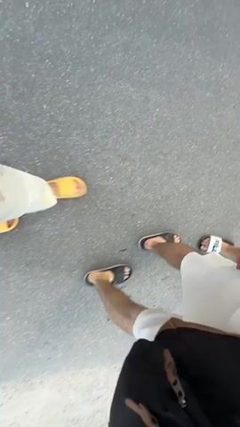 The video begins with a first-person perspective, showing feet wearing slides walking on a grey, asphalt-like surface. A person in yellow slides is visible on the left, and several other people in black and white slides are also visible. The camera pans slightly, showing more people walking. The scene then shifts to an outdoor environment. The camera is held by a person looking up and to their right, showing the sky and trees. Another person is visible in the foreground, facing away from the camera. In the distance, a pathway runs alongside a body of water, with several figures walking along it. The overall impression is of a group of people walking together outdoors, with some of the conversation possibly being about wealth.