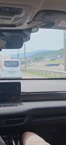 The video is filmed from the interior of a car, looking forward through the windshield. In the foreground, the dashboard and a car's infotainment screen are visible. In the mid-ground, a white van is driving on a road, followed by a large truck with a white cylindrical tank. The road appears to be an open highway with mountains and some buildings visible in the distance. The sky is overcast, suggesting a cloudy day. The car is moving, as indicated by the slight blur in the background.