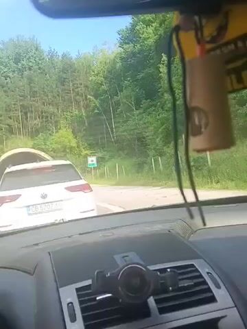 This video captures a view from inside a car as it approaches a tunnel. The scene is set on a sunny day, with lush green trees lining the road on either side, suggesting a rural or mountainous location. The road itself is asphalt, and there are clear lane markings.

The primary subjects are vehicles. In front of the car, a white Volkswagen station wagon with a Bulgarian license plate "CB 5707 AO" is stopped, waiting to enter a tunnel. To the left of the Volkswagen, a dark-colored minivan is also visible. The tunnel entrance is circular and appears to be carved into the mountainside, surrounded by trees. Two green road signs with white symbols are placed on either side of the road before the tunnel.

The activity taking place is vehicles queuing to enter a tunnel. The car from which the video is filmed is slowly approaching the tunnel entrance. The overall mood is calm, with the bright daylight and the natural surroundings creating a peaceful atmosphere.

There is visible text on the white Volkswagen's license plate and on a green and brown road sign to the left of the road, which appears to be in Bulgarian. The sign on the left side of the road, near the minivan, is a rectangular brown sign with text and a green border. Another green sign with a white symbol is visible on the right side of the road, near the tunnel entrance. The car's dashboard is also partially visible, showing gauges and a phone mount with a phone attached.