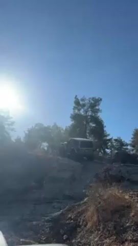 The video shows a gray SUV navigating a rocky slope, surrounded by trees, under a bright sun and blue sky. A hip-hop track plays in the background. The camera appears to be inside another vehicle that is stationary.