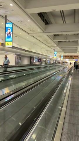 A person is walking on a moving walkway inside a brightly lit airport terminal. The walkway is flanked by another moving walkway going in the opposite direction. Above the walkways are suspended signs displaying advertisements. The terminal has a high ceiling with exposed structural elements and fluorescent lighting. The perspective of the video is from a first-person view, moving forward along the walkway. The overall environment appears clean and modern.