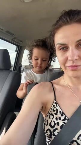 A woman is in the front passenger seat of a vehicle and films herself. A young child is in the backseat behind her and touches the back of her seat. The child is wearing a white t-shirt and green shorts. The woman is wearing a leopard print tank top and a necklace. The video is taken from inside the car, with the camera facing forward. Sunlight streams into the car through the windows. The woman turns the camera toward the child, who looks at the camera and opens their mouth. The woman then turns the camera back to herself.