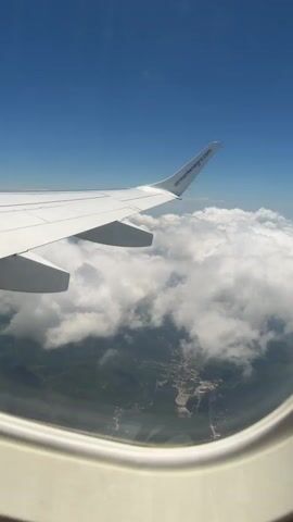 The video shows a view from an airplane window. The wing of the airplane is visible in the foreground, with the logo "airmontenegro.com" printed on the wingtip. Below the wing, a layer of white clouds stretches across the sky, obscuring much of the landscape below. Glimpses of green hills and a small town or settlement are visible through gaps in the clouds. The sky is a clear, bright blue. The camera appears to be panning slightly or the plane is in motion, causing a subtle shift in the perspective. The overall scene is one of aerial travel, with a focus on the view from the aircraft.