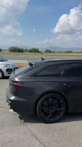 The video opens with a rear-view shot of a matte black Audi RS6 Avant stationary on a road. The car is parked on asphalt with white line markings. In the background, there is a highway, a grassy embankment, and distant hills under a blue sky with scattered clouds. The camera then pans slightly, revealing another car, a dark-colored SUV, to the left of the Audi. As the video progresses, more cars appear on the highway in the distance, including a dark hatchback. The Audi remains the primary focus for the initial part of the clip, with its distinctive rear spoiler and quad exhaust pipes visible. The environment appears to be a rural or semi-rural area, possibly alongside a highway construction zone or rest area, indicated by road signs and barriers. A tall green hedge lines the right side of the road, partially obscuring the view of the surroundings as the camera continues to move. The video ends with the Audi and other vehicles visible on the road, under the same cloudy sky.