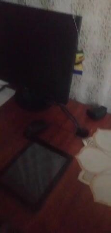 The video pans across a wooden desk. On the desk, there is a black computer monitor, a black mouse, a tablet, a small black object, a few paper napkins with floral designs, a piece of white paper with a pen, and a framed picture. The lighting is dim and the camera quality is low.