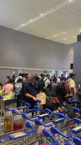 The video shows a busy airport check-in area. A long line of people, many with luggage carts, are waiting. The carts are predominantly blue and have multiple compartments for luggage. The people are diverse in age and gender. They are dressed casually, as one would expect for air travel. In the background, there are large digital screens displaying advertisements and flight information. The airport interior is modern, with high ceilings, large pillars, and a well-lit environment. Some individuals appear to be looking at their phones or conversing with each other while waiting. The overall atmosphere suggests a typical airport scene with significant passenger traffic.