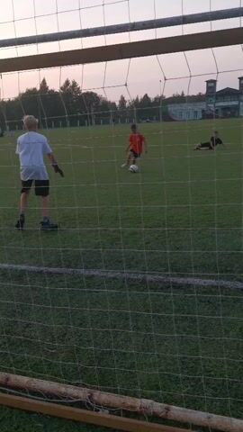 A group of children are playing soccer on a grassy field in front of a soccer goal. The camera is positioned behind the goal net, looking out onto the field. A child in a white shirt and black shorts is acting as the goalkeeper, standing in front of the goal. Two other children are on the field, one in an orange shirt and black shorts, and another in a black shirt. The child in the orange shirt is kicking a soccer ball towards the goal. The child in the black shirt is lying on the ground to the right of the field. In the background, there are trees, bleachers, and a building. The sky is overcast.