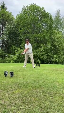 A young man is teeing off on a golf course. He is wearing a white polo shirt and beige pants with white shoes.  He swings the golf club and watches the ball as it flies forward. To his right are two distance markers indicating 61 and 57. The tee box is well-manicured green grass. Behind the tee box is a line of large, leafy trees. The sky is overcast.