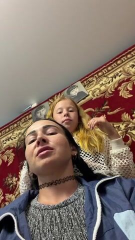 The video shows a woman and a young girl in an indoor setting, likely a home in Bronnitsa, Russia. The woman is in the foreground, facing the camera with a neutral expression. She has long, dark hair and is wearing a gray, speckled top and a blue and light blue jacket. A black choker necklace adorns her neck. Behind her, the girl with long, blonde hair is looking directly at the camera with wide, excited eyes. She is wearing a white, mesh-like top. The girl's hands are near the woman's head, and she appears to be braiding or styling the woman's hair. The background features a large, ornate red rug with a patterned design, which hangs on the wall. Two framed black and white photographs are also visible on the rug. The lighting suggests it is daytime. The overall mood appears to be playful and affectionate between the two individuals. There is no visible text in the video.