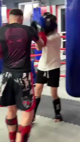 Two men spar in a gym. The man on the left is wearing red gloves, black and red shorts, a black shirt with a portrait on the back, and red foot protectors. He's punching the man on the right. The man on the right is wearing black gloves, a black helmet, a white t-shirt, black shorts, and black foot protectors. The gym has boxing rings in the background.