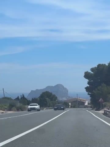 The video shows a drive on a paved two-lane road on a bright sunny day. There are cars passing by, including a white SUV, a black sedan, and a red car. The road has white lane markings.  In the distance, there is a large rock formation rising out of the water, partly obscured by a light haze. On the right side of the road, there is a house and a road sign depicting a bicycle. A tall pine tree is on the right side of the frame. Power lines stretch overhead. The sky is blue with some scattered white clouds. Music can be heard.