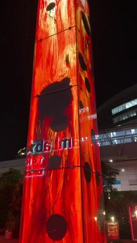 The video shows a tall, illuminated, rectangular structure at night. The structure is covered in a dynamic, abstract light display that appears to be in shades of red and orange, with flowing lines and dark circular shapes. The display has a glowing effect. The bottom section of the structure features text that is partially obscured and difficult to read clearly, but appears to be illuminated in neon-like colors. In the background, there are glimpses of buildings and trees, suggesting an urban or outdoor setting. The overall impression is that of a modern art installation or a prominent advertising display.