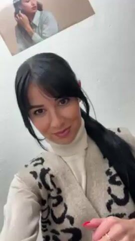 A woman with long black hair and bangs, wearing a white turtleneck and a tan cardigan with black leopard print, is standing in front of a white wall. She is looking at the camera with a slight smile. She blows a kiss towards the camera, then pushes a strand of hair behind her ear. A picture of a woman wearing jewelry is on the wall in the upper left of the frame. The woman has red nails.