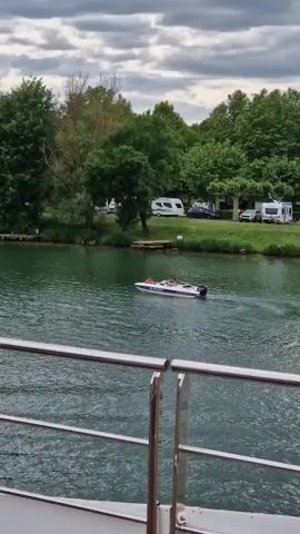 A white speedboat with a person onboard tows a small inflatable boat carrying three people on a green river. The speedboat's engine is on, causing a wake in the water. The inflatable boat has two people facing forward and one person facing backward. In the background, a green shoreline with trees and a campsite with several white trailers are visible under a cloudy sky. The video is filmed from a higher vantage point, possibly from another boat, with a metal railing visible in the foreground.