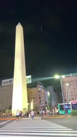The video shows the Obelisco de Buenos Aires at night, illuminated by bright lights. People are crossing the street on a crosswalk in front of the monument. There are several buildings visible in the background, with various commercial signs displayed on them. Traffic lights are also visible. The night sky is dark and clear.