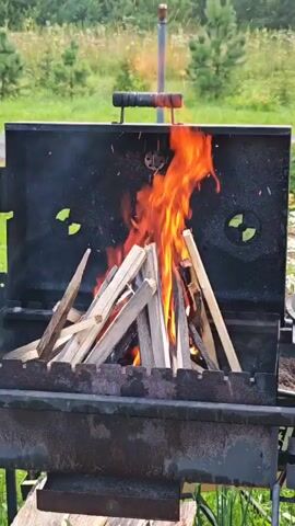 In Podosinki, Russia, a barbeque grill is shown with a fire burning in it. The grill is made of black metal and is filled with pieces of firewood. The flames are a bright orange and yellow, and there are sparks flying upwards. The background is a blur of green trees and grass, suggesting an outdoor setting. The time of day appears to be daytime, and the weather is likely warm enough for a barbeque. There are no people visible in the image, and no text is present. The overall impression is one of a pleasant, warm day in the countryside, with the anticipation of an outdoor meal.