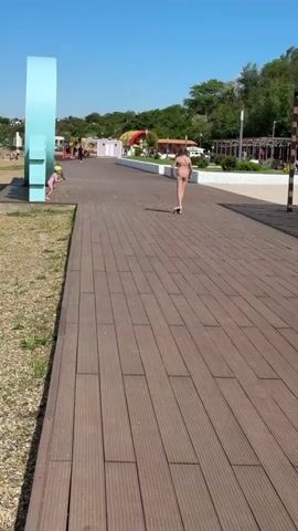 In a bright daylight scene in Taman, Russia, two children are active on a wooden boardwalk. An older girl, wearing a light-colored swimsuit, propels herself forward on a scooter. A younger child, wearing a pink outfit and a light-colored helmet, runs alongside or slightly behind her. The boardwalk is made of brown composite planks, stretching into the distance. To the left, a large, light-blue, circular archway or sculpture is visible. In the background, there's a grassy area with green trees, small white buildings (possibly changing rooms or kiosks), and other distant park structures under a clear blue sky. The children appear to be enjoying their time in this sunny recreational setting.