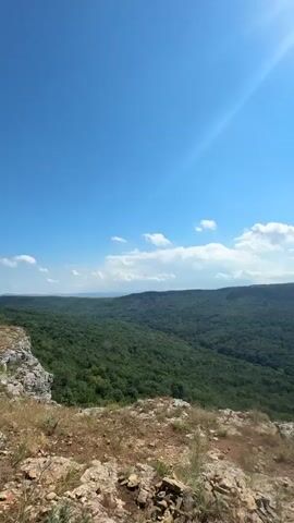 The video is a landscape view from a rocky cliff overlooking a vast, dense forest under a bright blue sky with scattered clouds and a shining sun. The foreground shows the rocky, uneven terrain of the cliff. The middle ground displays an expansive green forest that stretches to the horizon. The background is dominated by the sky with the sun creating a lens flare effect. There are no people clearly visible or performing any actions, although a hand with a black scrunchie is visible on the right side of the frame in the last two seconds. The environment appears to be natural and rural.
