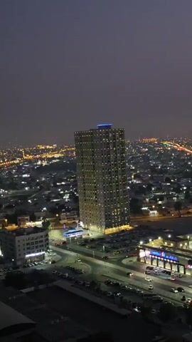 This is an aerial video capturing a cityscape at night, likely in Ajman, United Arab Emirates. The prominent subject is a tall, modern apartment building illuminated with rows of green lights, giving it a striking appearance against the dark sky. The building's top is crowned with blue and pink lights.

The surrounding area is a sprawling urban landscape dotted with numerous lights, indicating a densely populated city. We can see a network of roads with cars moving, suggesting ongoing activity. A large parking lot is visible near the apartment building, filled with vehicles. In the foreground, another building with illuminated windows and a sign for "Carrefour" is clearly visible, indicating a shopping center or hypermarket. The overall scene suggests a vibrant urban environment at night.

The time of day is clearly night, with the sky being dark and artificial lights being the primary source of illumination. The weather appears clear, with no visible signs of rain or fog. The presence of a Carrefour store strongly suggests an urban or suburban retail hub. The mood conveyed by the video is one of dynamism and urban life continuing into the night.