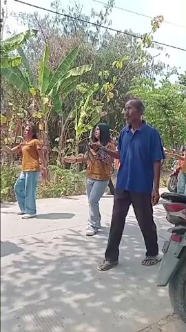 A group of young women in orange shirts and jeans are dancing in a line on a paved road. An older man in a blue shirt and dark pants walks in front of them, looking to his left. Behind the dancers, there are trees and buildings. A motorcycle is parked to the right of the frame. The scene appears to be outdoors during the day.