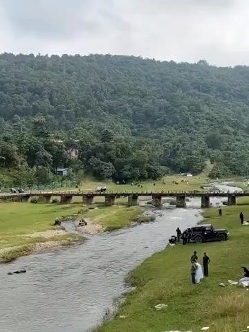 A wide shot captures a natural landscape with a river flowing in the foreground. On the right bank of the river, several people are gathered near two parked vehicles, one of which is a black SUV. Further to the right, there are a few more individuals and some scattered items. The river is spanned by a concrete bridge with multiple arches, allowing a grassy area with scattered trees and more people to be visible on the other side. Beyond the bridge, a lush, green, tree-covered hill forms the backdrop, with a cloudy sky overhead.