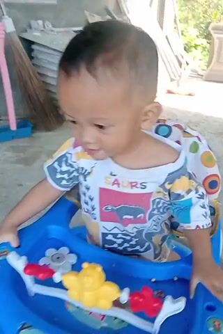 A toddler stands in a blue baby walker. The walker has a tray with toys attached, including a yellow bear and red objects. The toddler is wearing a white t-shirt with colorful patterns and dinosaur graphics. The toddler is looking down at the toys. The background appears to be an outdoor or semi-outdoor area with a concrete floor and some natural light.