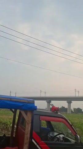 Here is a concise description of the video:

The video shows an elevated road or bridge with concrete pillars, spanning across a grassy area. Power lines run parallel to the road or bridge. The sky is overcast with a hint of light suggesting either sunrise or sunset. As the video progresses, the camera pans slightly to the right to reveal a road adjacent to the grassy area. A white van drives away from the camera on the road. The road appears somewhat cracked and worn. There are trees lining the right side of the road. A voice in the background mentions numbers.