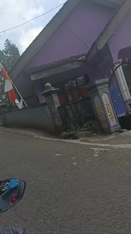 The video shows a damaged house from a ground-level perspective, possibly from a moving vehicle. The roof of the house has collapsed and is tilted at a severe angle. Sections of the wall appear to be missing or broken, revealing the interior. Debris is scattered on the ground in front of the house. The surrounding environment appears to be rural, with trees and vegetation visible in the background. The sky is cloudy. There are no people clearly visible in the video.